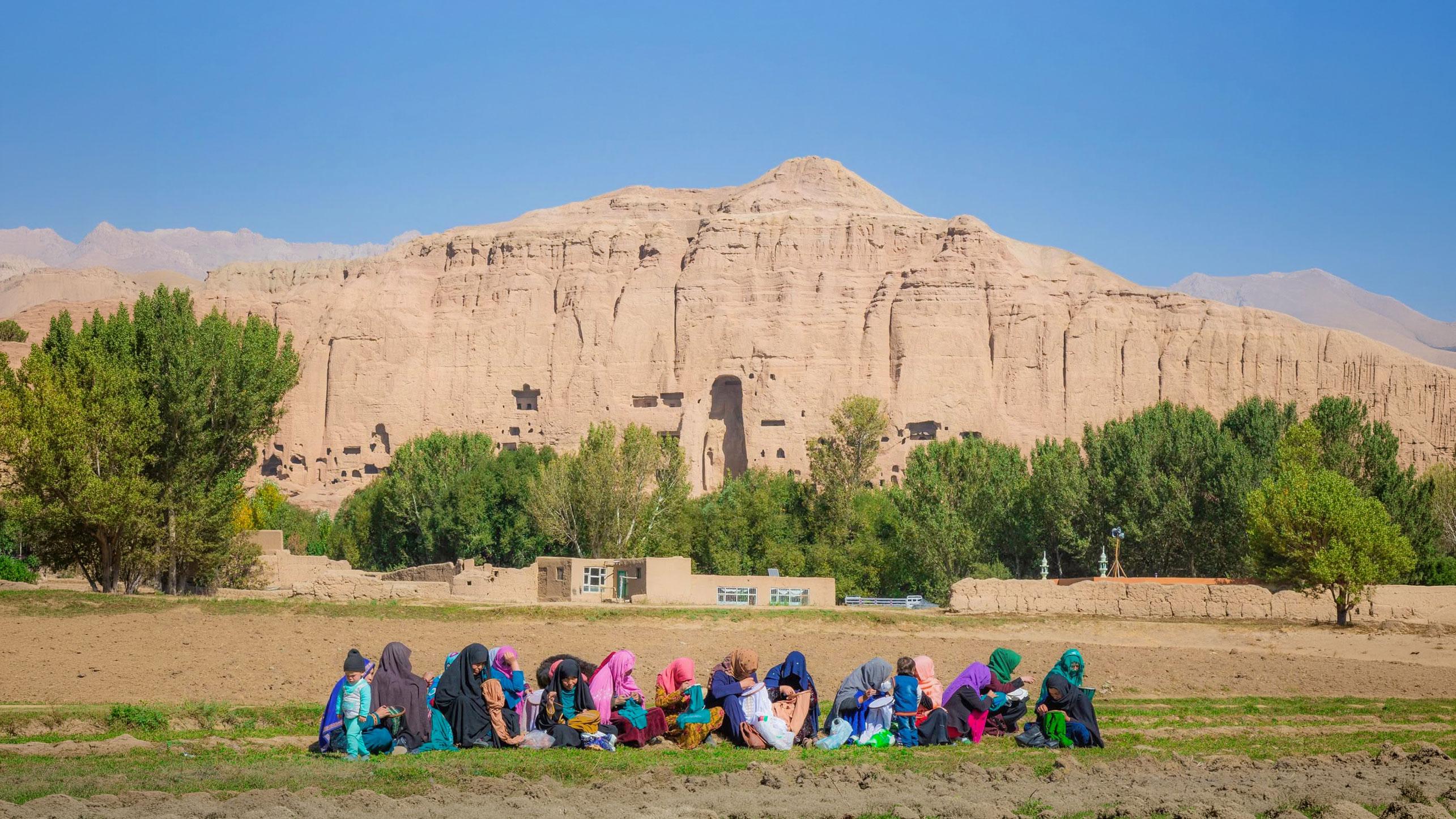 A group of women sitting together in a field doing embroidery, with cliffs and trees behind them.