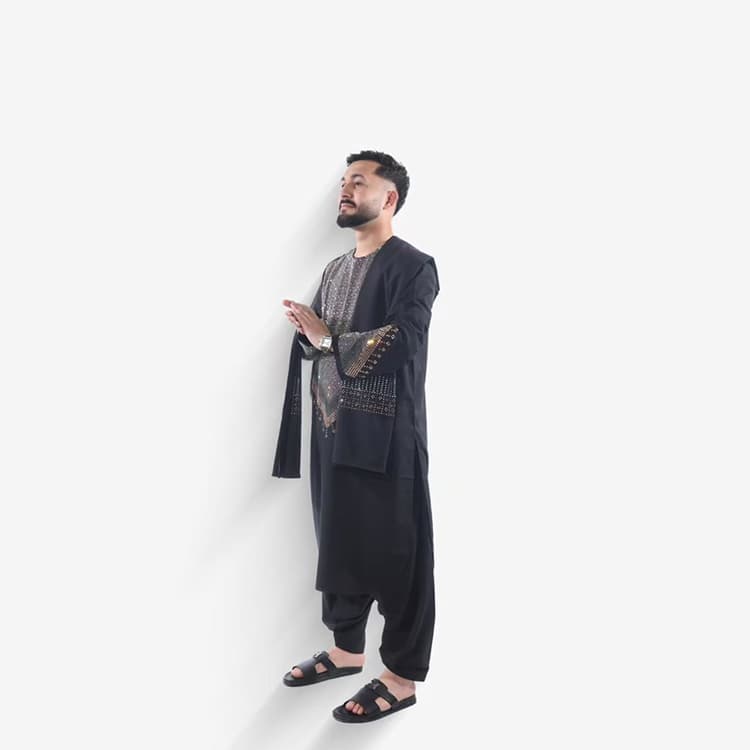 A man wearing a long black tunic and loose trousers with gold embroidery at the chest and cuff.