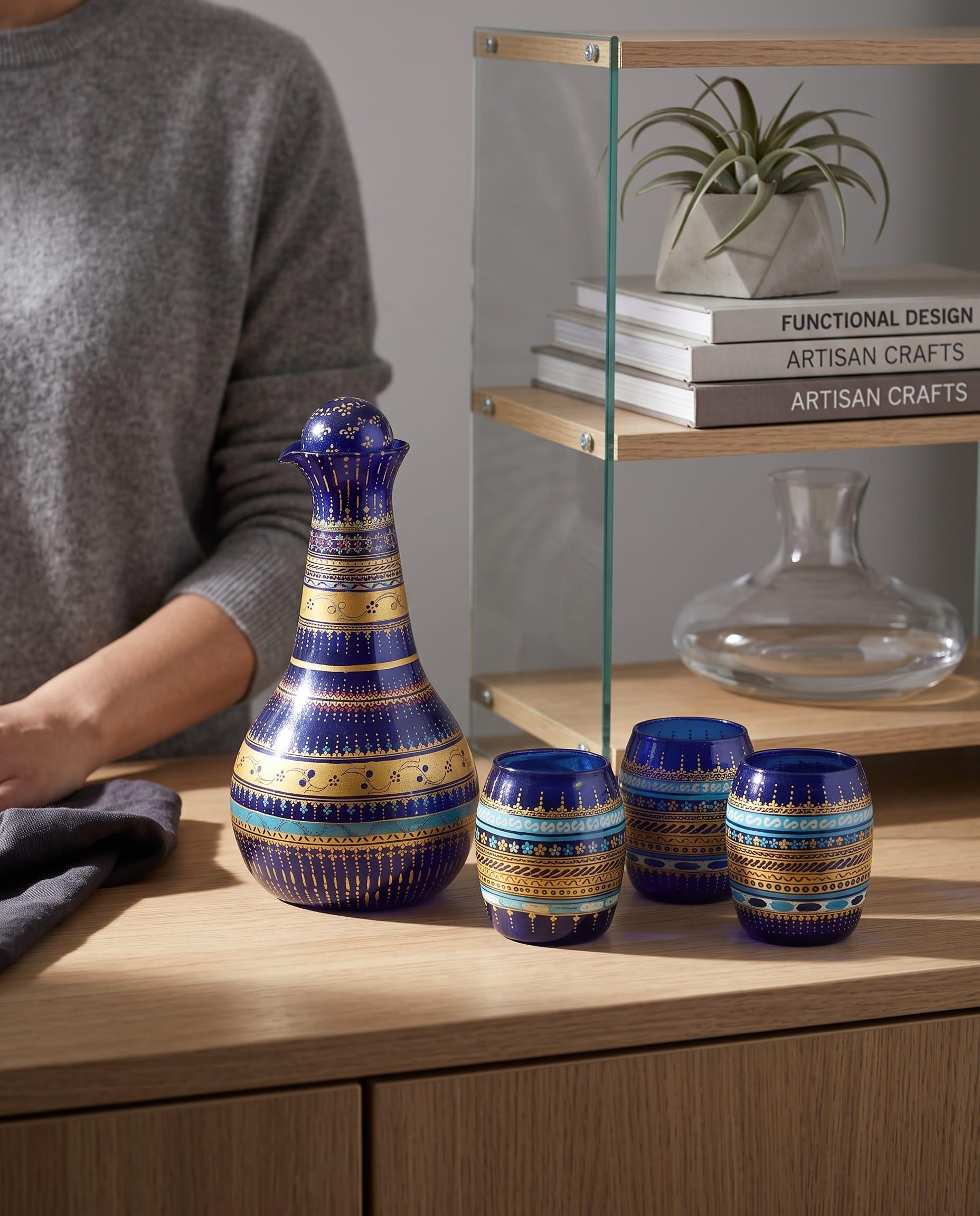 A carafe striped in blue and gold with three matching cups on a wooden table.